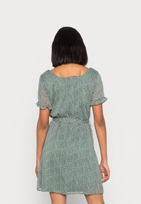 Vestido verde de manga corta con cintura ajustada y mangas con volantes. La tela presenta lunares blancos y una falda fluida. Se muestra la vista posterior.