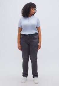 Ανοιχτό μπλε πλεκτό πόλο πουκάμισο με γιακά, συνδυασμένο με σκούρο γκρι τζιν και λευκά αθλητικά παπούτσια, σε όρθια στάση μπροστά σε απλό φόντο.