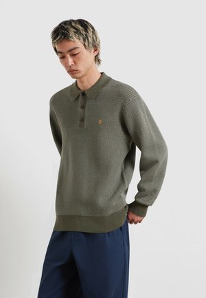 Giovane uomo con capelli biondi corti che indossa un maglione polo lavorato a maglia con motivo verde oliva e pantaloni blu scuro, guarda verso il basso con le mani dietro la schiena.