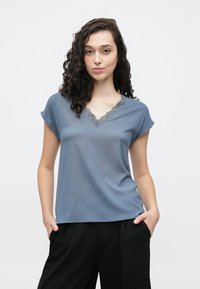 Sininen, kevyt, lyhythihainen paita V-päällä ja pitsireunuksella. Sileä pinta, rento istuvuus, hienovarainen laskeutuvuus, yhdistetty mustiin housuihin.