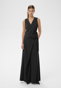 Vrouw staat in een mouwloze zwart wikkelvest en wijde zwarte broek met een riem voor een effen witte achtergrond.