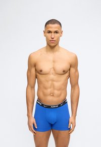 Γυμνασμένος άντρας στέκεται φορώντας μπλε ανδρικά εσώρουχα Nike με μαύρη μέση, σε λευκό φόντο.