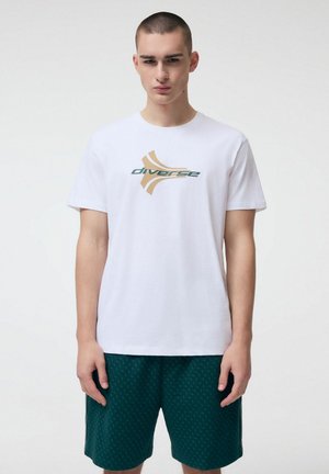 Młody mężczyzna z krótkimi włosami, ubrany w białą koszulkę z logo "diverse" i ciemnozielone krótkie spodenki w wzory, stoi na jednolitym tle.