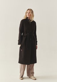 Sort uldfrakke med høj krave, to brune knapper og et matchende bælte. Båret over lysebrune vide bukser og beige sneakers.