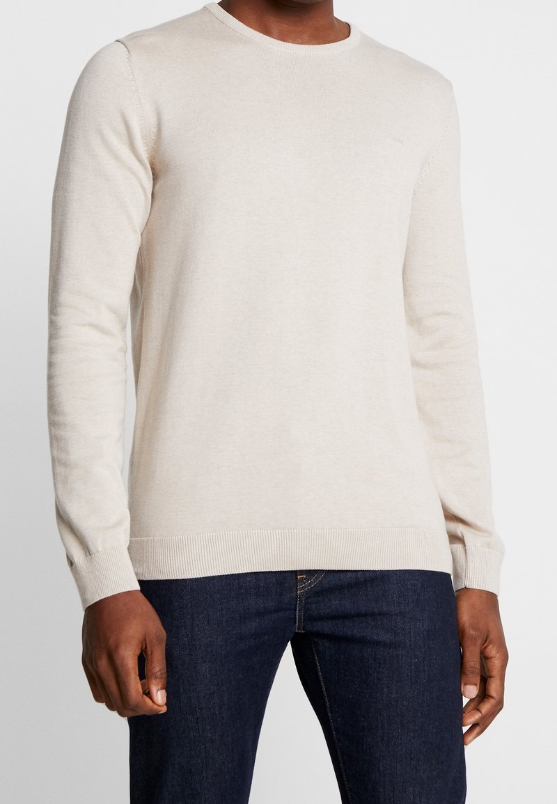 Homme portant un pull uni beige clair à manches longues associé à un jean denim bleu foncé, debout devant un fond blanc.