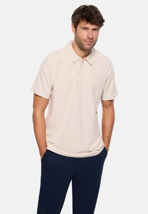 Hombre con camisa tipo polo de manga corta beige y pantalones azul marino, de pie con las manos en los bolsillos contra un fondo liso.