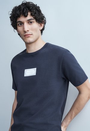 Νέος άνδρας με σκούρα σγουρά μαλλιά, φορώντας μπλε σκούρο μπλουζάκι με μικρό λευκό ορθογώνιο λογότυπο, σε ανοιχτό γκρι φόντο.