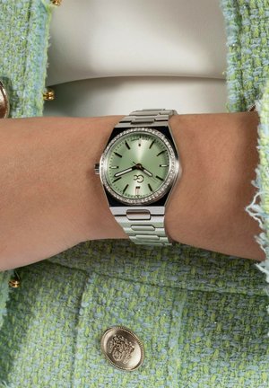 Reloj de metal plateado con una esfera verde menta, marcadores de hora texturizados y acentos de cristal. Lleva puesto en una muñeca con un tejido ligero verde texturizado.