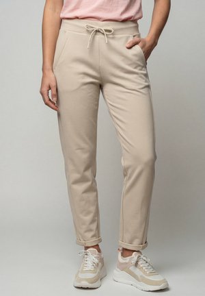 Person trägt beige Kordelzughose mit hochgekrempelten Bündchen und beige-weißen Turnschuhen, steht mit einer Hand in der Tasche.