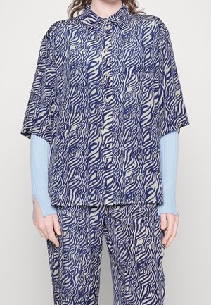 Personne portant une chemise et un pantalon amples assortis avec un motif abstrait ondulé bleu et beige et des manches longues côtelées bleu clair visibles sous la chemise.