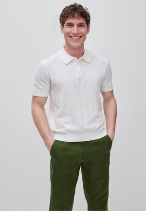 Lachende man met krullend bruin haar, gekleed in een gestructureerd wit poloshirt met korte mouwen en olijfgroene broek, staand met handen in de zakken.