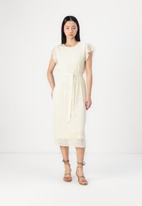 Robe midi en dentelle blanche avec des manches courtes à volants, une taille cintrée à nouer, et un ourlet festonné, associée à des sandales à lanières beiges.