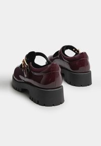 Zapatos de charol burdeos con un diseño de corte, herrajes dorados y suelas de goma negra gruesa con un patrón de tacos.