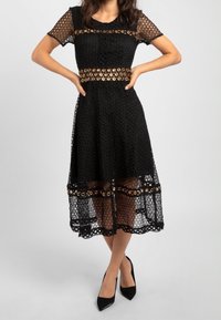 Robe noire en crochet avec manches courtes, découpes circulaires et ourlet mi-long. Détails avec une taille à motifs et une bordure en dentelle.