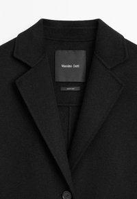 Černý vlněný kabát s klopovým límcem, zapínáním na jeden knoflík a měkkou texturou. Štítek uvnitř říká "Massimo Dutti, vyrobeno v Číně."