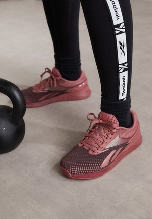 Nohy obuté v růžových teniskách Reebok a černé legíny s logem Reebok stojí vedle černé kettlebell na podlaze posilovny.