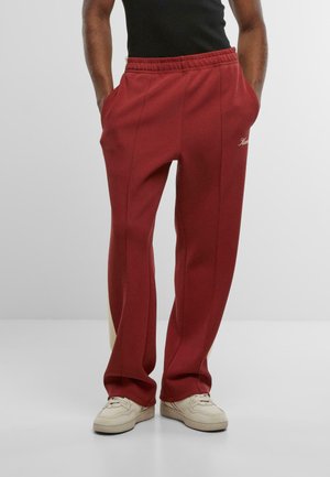 Person, der rote Sporthosen mit Seitentaschen und weiße Sneakers trägt, steht mit den Händen in den Taschen vor einem schlichten Hintergrund.