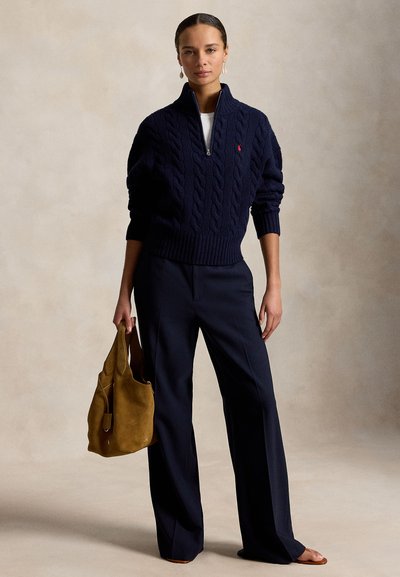 Nő, aki tengerészkék, kötött fél cipzáras pulóvert és bőszárú nadrágot visel, barna velúr táskát tart, semleges háttér előtt áll.