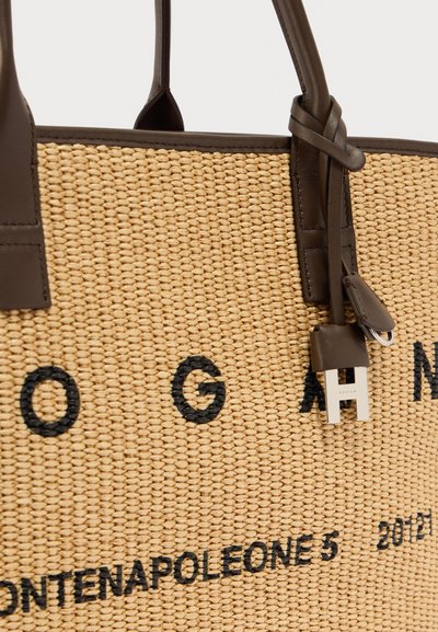 Sac fourre-tout tissé beige avec des poignées en cuir marron foncé, texte imprimé en noir, et une étiquette en cuir avec un pendentif argenté en forme de "H" attaché.