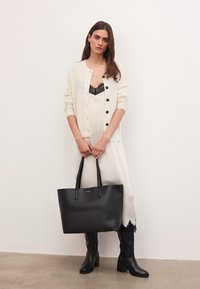 Kvinna i vit klänning med svart spetsdetalj, krämfärgad cardigan med knappar, svarta knä höga stövlar och bär en stor svart tote-väska.
