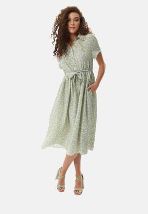 Button-up, short-sleeve dress in light green fabric with floral print, cinched waist and belt, featuring side pockets. Paired with green heels.