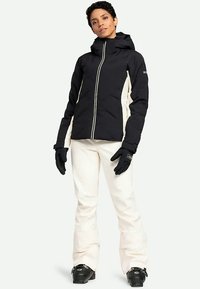 Persona che indossa una giacca da sci bianca e nera, pantaloni da neve bianchi, guanti neri e scarponi da sci neri, in piedi contro uno sfondo semplice.