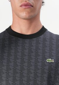 Hanorac negru cu model, având un design geometric, guler ribit negru și un mic logo verde de crocodil pe piept.