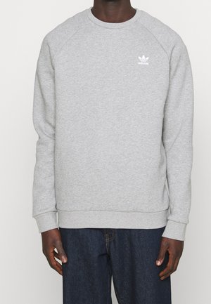 Man in een lichtgrijze Adidas crewneck sweatshirt met lange mouwen en een donkere spijkerbroek, staande tegen een effen achtergrond.