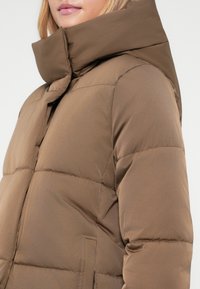 Casaco puffer castanho com colarinho, acabamento liso, fecho éclair na frente e acolchoamento proeminente. Possui bolsos laterais e um corte descontraído.