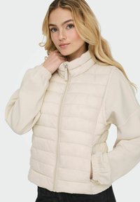 Hellbeige gesteppte Weste mit hohem Kragen, durchgehendem Reißverschluss, elastischen Bündchen und Seitentaschen; weiche Textur mit gesteppten horizontalen Linien.