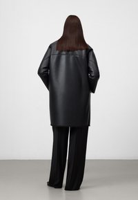 Zwarte faux leren oversized jas met verlaagde schouders, een soepele textuur en een knielange uitstraling, gecombineerd met zwarte wijde broekspijpen.