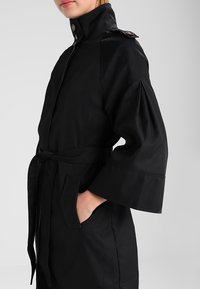 Černý trenčkot s vysokým límcem, stahovaným pasem a širokými rukávy. Obsahuje pásek, kapsy a knoflíkové detaily.