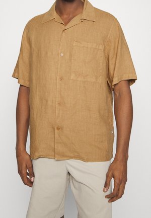 Hombre con camisa de manga corta color marrón claro con bolsillo en el pecho y pantalones cortos beige claro frente a un fondo liso.