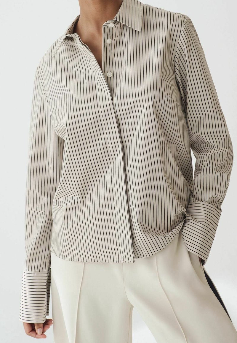 Personne portant une chemise beige à manches longues avec des fines rayures noires verticales et un pantalon blanc, une main dans la poche, sur un fond uni.