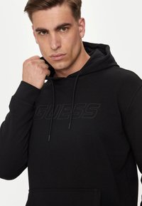 Hoodie nero realizzato in morbido tessuto, dotato di cappuccio con cordino, tasca a marsupio anteriore e logo "GUESS" in rilievo sul petto.