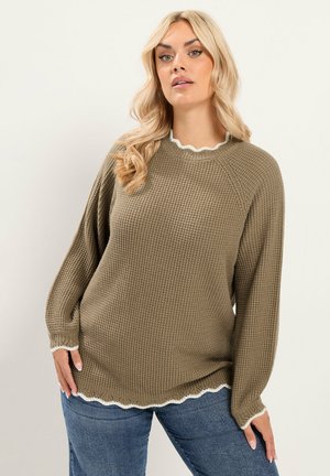 Donna bionda che indossa un maglione a maglia marrone largo con bordo bianco smerlato su collo, polsini e orlo, abbinato a jeans blu.