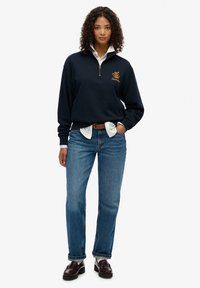 Navyblauwe pullover sweatshirt met hoge kraag en kwartrits, met geborduurd logo. Gecombineerd met lichtblauwe rechte jeans en donkere instappers.