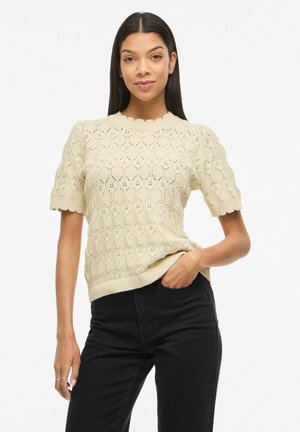 Femme portant un haut en maille crème à manches courtes avec un col festonné et un pantalon noir, debout avec une main dans la poche.