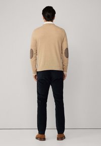 Bežani pulover s laktovnim oblogama, uparen s tamnim hlačama i smešnim cipelama, prikazan na osobi koja je okrenuta leđima. Glatka tekstura, jednostavan dizajn.