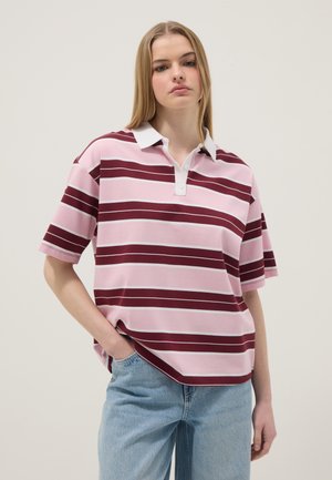 Jong persoon draagt een roze en bordeauxrood gestreept poloshirt en lichtblauwe spijkerbroek, staand met één hand in de zak tegen een neutrale achtergrond.