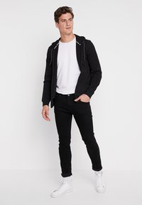 Zwarte zip-up hoodie met witte lange trekkoorden, gecombineerd met een wit t-shirt en zwarte spijkerbroek. Witte sneakers maken de look compleet.