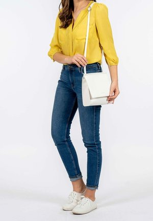 Kobieta ubrana w żółtą bluzkę, niebieskie dżinsy, białe tenisówki i nosząca białą torebkę na ramię z klapą.