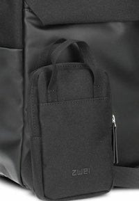 Piccola borsa nera in tessuto con doppi manici e cerniera, attaccata a una borsa nera più grande con materiali lisci e testurizzati.