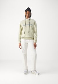 Světle zelená mikina s modrým logem "Carhartt", dlouhé rukávy a šňůrka. Spárovaná s bílými volnými kalhotami a šedými teniskami.