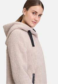 Cappotto con cappuccio rosa chiaro realizzato in tessuto strutturato, con accessori neri sulle coulisse e una tasca con zip sul lato.