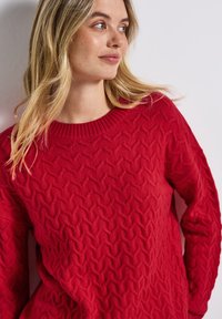 Roter gestrickter Pullover mit einem strukturierten Zopfmuster und einem gerippten Kragen. Lange Ärmel und lockere Passform. Weicher, kuscheliger Stoff.