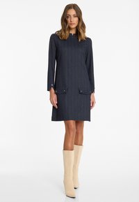 Gestreiftes navy Kleid mit hohem Kragen, langen Ärmeln und frontalen Taschen, aus strukturierter Stoff gefertigt. Tragen mit hohen beigen Wildlederstiefeln.