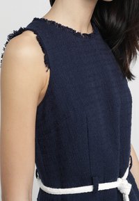 Námořnická modrá bezramínková šaty s třepenými okraji, strukturovanou látkou a bílým uvázaným pásem v pase. Minimalistický design s přiléhavým střihem.