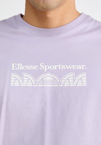 Gaišs lavandas krāsas kokvilnas T-krekls ar baltu "Ellesse Sportswear" logo, kas ar izliekta strīpu dizainu izvietots šķiņķī. Klasisks apaļais kakls.