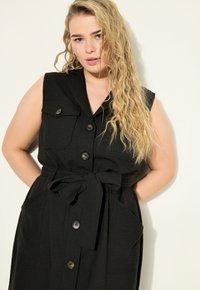 Abito nero senza maniche con colletto, chiusura frontale a bottoni, due tasche sul petto e una cintura in vita per una vestibilità aderente. Tessuto testurizzato.